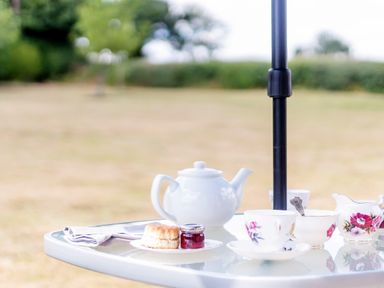 A glass table with a teapot scone jar of jam and teacups outdoors at Norbank Garden Studio in Bressingham