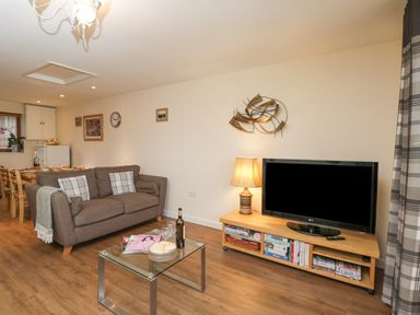 A living room with a brown sofa a glass coffee table with plates glasses and a wine bottle a wooden TV stand with a TV and a lamp at Roan Cottage in Royal Wootton Bassett