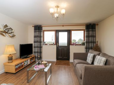 A living room with a brown sofa and checkered pillows a glass coffee table wooden tv stand with a lamp a television and wall art at Roan Cottage in Royal Wootton Bassett