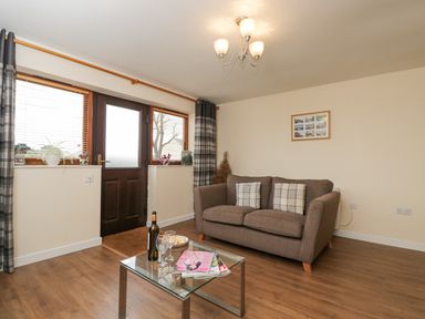 A living room with a brown sofa and glass coffee table near a door with two windows at Roan Cottage in Royal Wootton Bassett