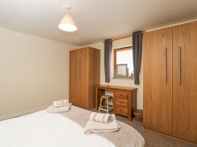 A bedroom with a bed, two wooden wardrobes, a wooden dressing table with a stool, a small window with curtains and towels on the bed at Roan Cottage in Royal Wootton Bassett