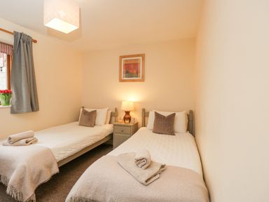 A bedroom with two single beds with beige blankets beside a small nightstand with a lamp and a framed picture on the wall at Roan Cottage in Royal Wootton Bassett