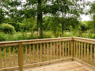 A wooden deck railing overlooking a forested area with trees and bushes at Chaffinch Lodge in Highampton near Sheepwash