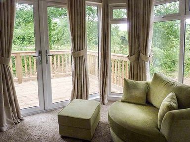 A corner room with green rounded sofa and ottoman in front of glass doors and windows at Chaffinch Lodge in Highampton near Sheepwash