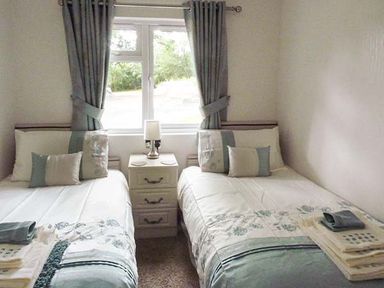 A bedroom with two single beds separated by a nightstand with a lamp and window with curtains at Chaffinch Lodge in Highampton near Sheepwash