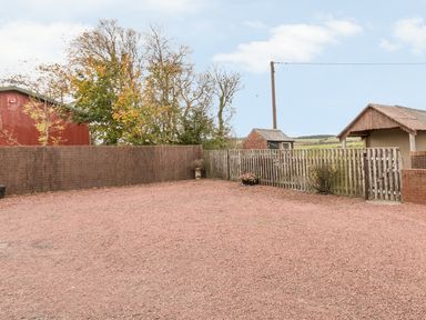 An outdoor gravel area enclosed by wooden fences with trees and small buildings in the background at St Cuthbert's Cottage in Beal near Holy Island