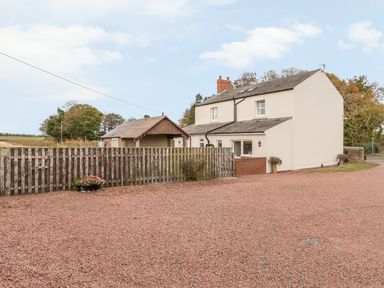 An outdoor view of a two-story house with a fenced yard and gravel driveway at St Cuthberts Cottage in Beal near Holy Island