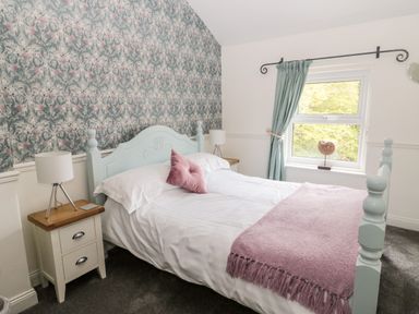 A bedroom with a double bed, bedside tables with lamps, floral wallpaper, and a window with curtains at St Cuthberts Cottage in Beal near Holy Island