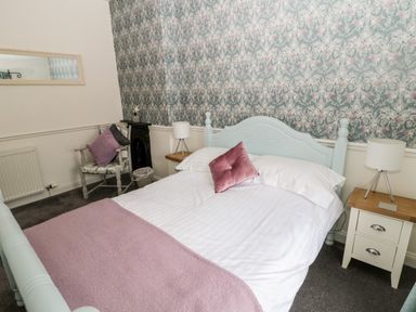 A bedroom with a white bed, bedside tables with lamps, a chair with cushions, and floral wallpaper at St Cuthberts Cottage Beal near Holy Island