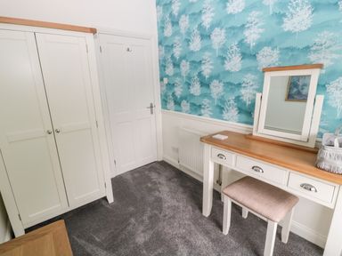 A room with a wardrobe a door a dressing table with mirror and stool and blue wallpaper with white tree design at St Cuthberts Cottage in Beal near Holy Island