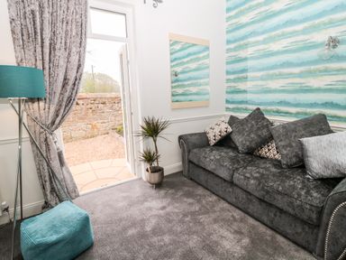 A room with a grey sofa with cushions a turquoise floor lamp a matching pouf and an open door leading outside at St Cuthberts Cottage Beal near Holy Island