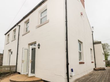 The exterior of a two story house with multiple windows a door a small patio area and a wooden deck at St Cuthberts Cottage in Beal near Holy Island
