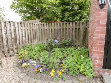 A corner garden bed with various green plants and colorful flowers beside a brick wall and wooden fence at St Cuthberts Cottage Beal near Holy Island