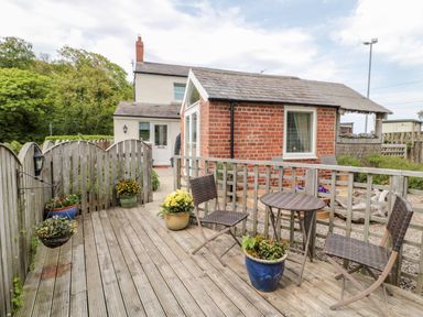 A wooden deck with chairs and a table surrounded by flower pots near a brick building at St Cuthberts Cottage Beal near Holy Island