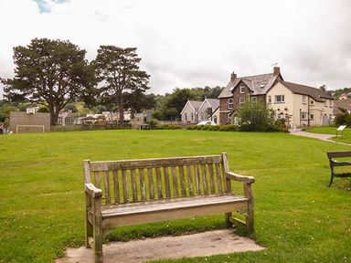 A wooden bench on green grass in a park with trees houses and a basketball hoop in the background at Rose Cottage in St Dogmaels near Cardigan