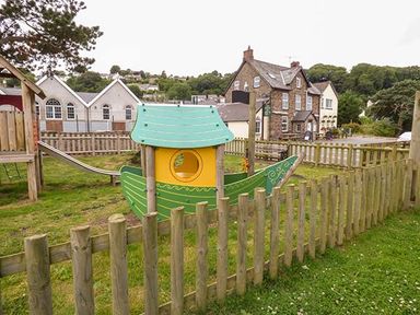 A fenced playground with a boat-shaped play structure and slide near stone and white buildings at Rose Cottage in St Dogmaels near Cardigan