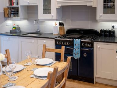 A kitchen with a wooden dining table set with plates and glasses a black oven with a blue oven mitt white cabinets and black countertop at Rose Cottage in St Dogmaels near Cardigan
