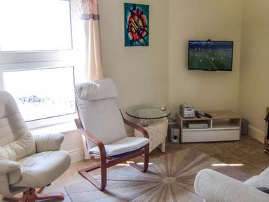 A living room with two armchairs a round glass side table a wall-mounted television and a wooden media unit at The Beach House Apartment in Buncrana County Donegal