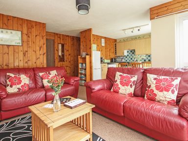 A living room with red leather sofas and floral cushions a wooden coffee table and a kitchen area at 4 Manorcombe in Honicombe Manor near Gunnislake