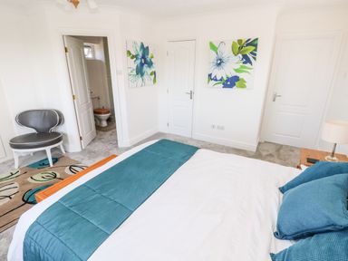 A bedroom with a bed covered with a teal runner and pillows a gray chair a patterned rug two floral paintings two closed white doors and an open door revealing a bathroom at 1 Atlantic Haven in Llangennith
