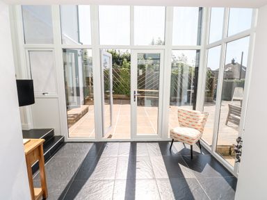 A room with large glass windows and doors overlooking a patio with a small chair and a wooden side table at 1 Atlantic Haven in Llangennith