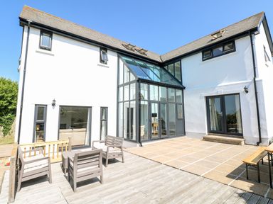 An outdoor patio area with wooden deck chairs and a table next to a white two-story house with large glass windows and doors at 1 Atlantic Haven in Llangennith