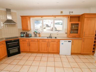A kitchen with wooden cabinets a double sink a dishwasher and a window showing parked cars at 1 Atlantic Haven in Llangennith