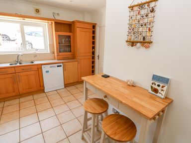 A kitchen with wooden cabinets a dishwasher a sink a small wooden table with two stools and a shell decoration on the wall at 1 Atlantic Haven in Llangennith