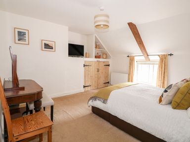 A bedroom with a bed, wooden chair, desk, framed pictures, TV, wooden doors, window, and exposed wooden beam at Flint Cottage in Swaffham