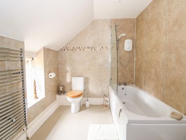 A bathroom with a bathtub shower combination a toilet with wooden seat a window with a lighthouse decoration and a towel radiator at Flint Cottage in Swaffham