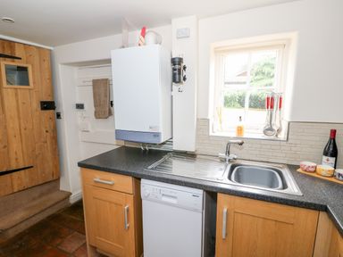 A kitchen with a wooden door tiled floor a white boiler above black countertop a sink under a window and wooden cabinets at Flint Cottage in Swaffham