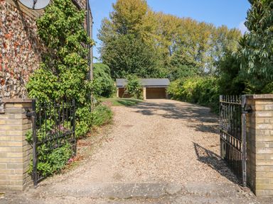A gravel driveway with open metal gates and a peg house wall with a climbing plant leading to a double garage with trees in the background at Flint Cottage in Swaffham