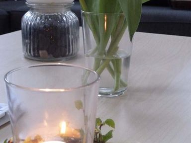A table with a lit candle in a glass holder a glass jar with a white lid and a glass vase with green stems at Capel Dinorwig in Llanberis
