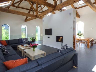 A living room with large dark sofas a coffee table with flowers a TV mounted on a wall and exposed wooden ceiling beams at Capel Dinorwig in Llanberis