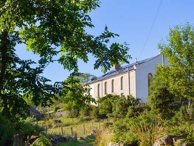 A white building with arched windows surrounded by trees and plants at Capel Dinorwig in Llanberis