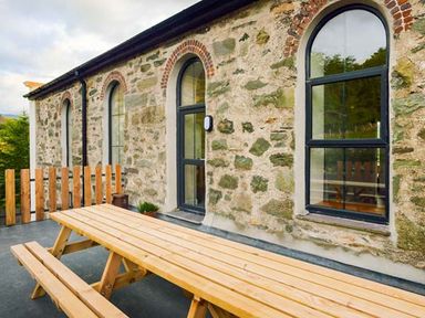 An outdoor area with a wooden picnic table and benches next to a stone building with arched windows at Capel Dinorwig in Llanberis