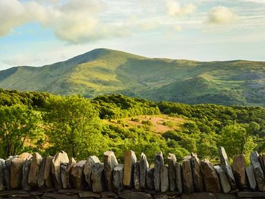 A stone wall in front of green trees and hills under a partly cloudy sky at Capel Dinorwig in Llanberis