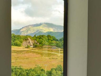 A view of green hills and mountains with power lines and trees seen through an open window at Capel Dinorwig in Llanberis