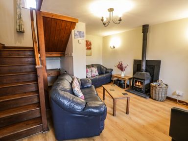 A living room with black leather sofas a wooden coffee table a wood burning stove and wooden stairs at Bwthyn Fedwr Gog in Maerdy near Bala