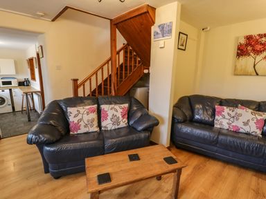 A living room with two black sofas with floral cushions a wooden coffee table and a wooden staircase at Bwthyn Fed'wr Gog in Maerdy near Bala