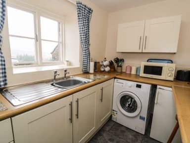 A kitchen with a sink under a window with curtains a washing machine microwave and various utensils on the countertop at Bwthyn Fed'wr Gog in Maerdy near Bala