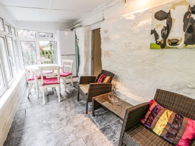 A room with white dining table and chairs beside a fridge and wicker chairs with colorful cushions at Bwthyn Fed'wr Gog in Maerdy near Bala