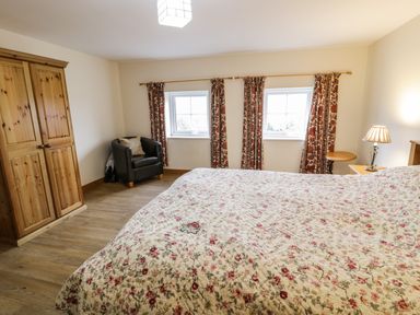 A bedroom with a floral bedspread a wooden wardrobe a black armchair two windows with floral curtains and a bedside table lamp at Bwthyn Fedwr Gog in Maerdy near Bala