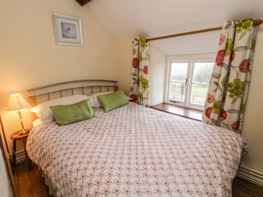 A bedroom with a double bed green pillows a bedside table with a lamp floral curtains and a window at Bwthyn Fed'wr Gog Maerdy near Bala