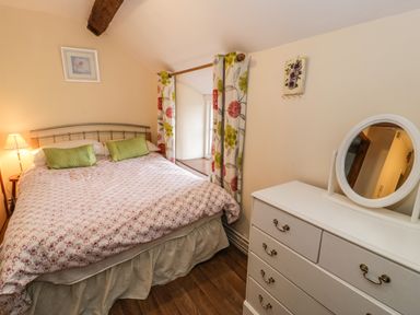 A bedroom with a double bed green pillows a bedside lamp floral curtains and a white dresser with a round mirror at Bwthyn Fedwr Gog Maerdy near Bala
