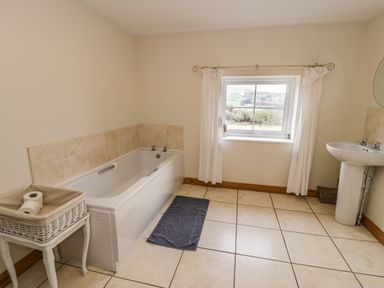 A bathroom with a bathtub beside a basket on a small table a window with curtains and a pedestal sink at Bwthyn Fedwr Gog in Maerdy near Bala