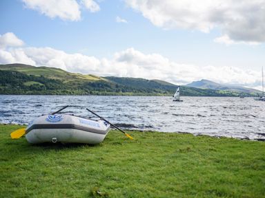 An inflatable boat with oars on grassy shore by a lake with hills and sailboats in the background at Bwthyn Fed'wr Gog Maerdy near Bala