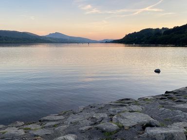 A rocky lakeshore with calm water and mountains in the background at Bwthyn Fed'wr Gog Maerdy near Bala
