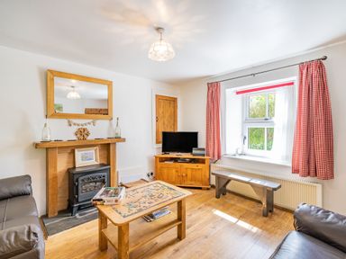 A living room with a wooden coffee table and puzzle pieces a fireplace with a large mirror and two bottles a television on a wooden stand a window with red checked curtains and a wooden bench at No 7 Spark Bridge near Ulverston