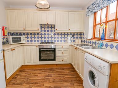 A kitchen with cream cabinets a microwave an oven a washing machine and a window with a blue vase of flowers at Little Venice in Whitby